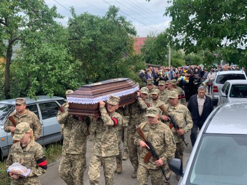 Military funeral procession with uniformed soldiers carrying a dark wooden coffin draped in a cloth, flanked by civilians and parked cars on a tree-lined street.