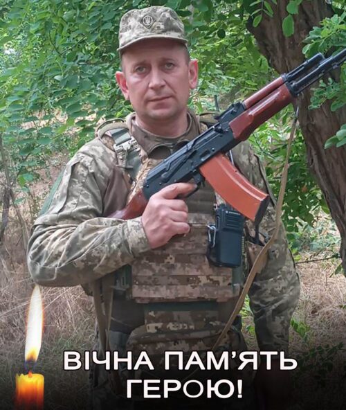 Soldier in camouflage holding a rifle outdoors with a candle and Ukrainian text reading 'Eternal memory to the hero' (Вічна пам'ять герою).