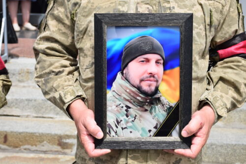 Soldier in camouflage uniform holding a framed photo of a man wearing a black beanie, with a Ukrainian flag visible in the background.