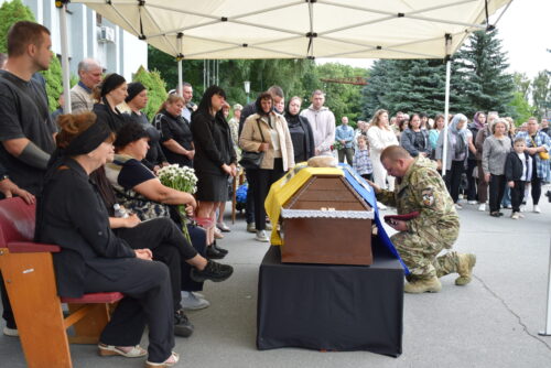 Mourning crowd under a white canopy gathers around a flag-draped coffin with a kneeling soldier praying beside it.