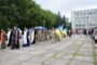 Military parade: Ukrainian soldiers march with blue and yellow flags, flanked by Orthodox priests in ceremonial robes, as spectators line a paved plaza.