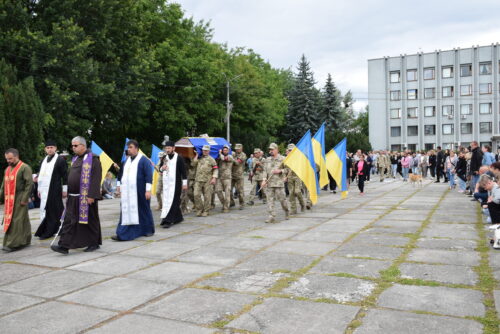 Military parade: Ukrainian soldiers march with blue and yellow flags, flanked by Orthodox priests in ceremonial robes, as spectators line a paved plaza.