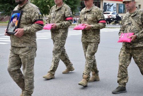 Soldiers in camouflage march down a street, one carrying a framed portrait of a bearded man while the others hold pink cushions or gifts, all wearing pink armbands/gloves as a solemn procession.