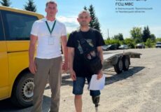Two men stand beside a yellow bus on a sunny day; one wears a white shirt with a lanyard, the other has a prosthetic leg and a black shirt, holding papers near a trailer, with a regional service center logo in the skyward corner.