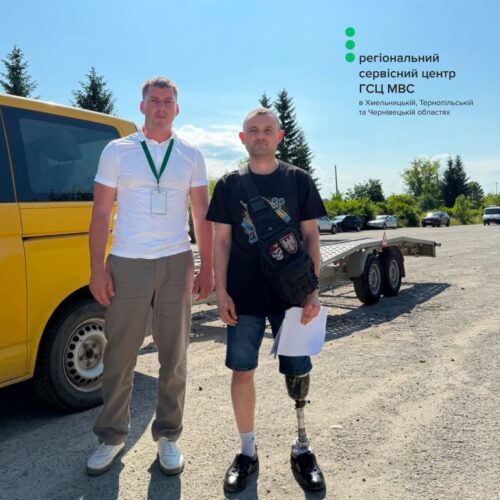 Two men stand beside a yellow bus on a sunny day; one wears a white shirt with a lanyard, the other has a prosthetic leg and a black shirt, holding papers near a trailer, with a regional service center logo in the skyward corner.