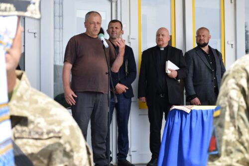 Man speaks into a microphone at a ceremony on a doorway porch, with four men in suits behind him and soldiers visible in the foreground.
