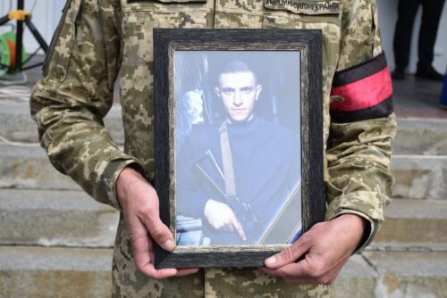 Soldier in camouflage holding a framed portrait of a man in ceremonial attire with a sword, standing on concrete steps.