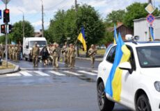 Ukrainian soldiers marching across a street with blue-and-yellow flags; a white SUV in the foreground draped with a Ukrainian flag.