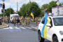 Ukrainian soldiers marching across a street with blue-and-yellow flags; a white SUV in the foreground draped with a Ukrainian flag.