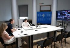 Three people sit at a white conference table with microphones in a hybrid meeting; a video call grid is displayed on a screen to the right.