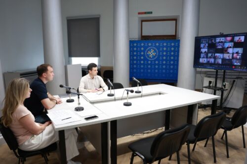 Three people sit at a white conference table with microphones in a hybrid meeting; a video call grid is displayed on a screen to the right.