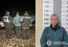 Two police officers in dark uniforms stand with a handcuffed man at night on a cobblestone surface; their backs are to the camera.