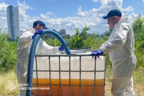 Two workers in hazmat suits handle a large IBC with orange liquid outdoors; emergency services emblem in the corner.