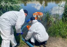 Three emergency responders in white hazmat suits work by a riverbank, handling a blue pipe for water safety operations near tall reeds in a calm lake.