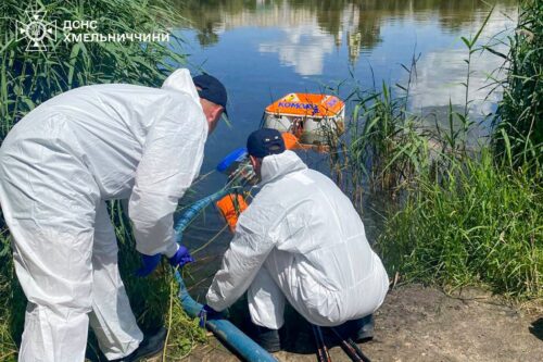 Three emergency responders in white hazmat suits work by a riverbank, handling a blue pipe for water safety operations near tall reeds in a calm lake.