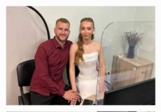 Couple posing in a photo studio: man in a maroon shirt and woman in a white strapless dress sit at a desk with a computer, backdrop visible behind them and a divider between them.