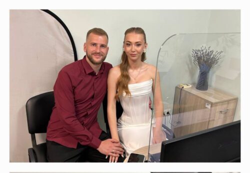 Couple posing in a photo studio: man in a maroon shirt and woman in a white strapless dress sit at a desk with a computer, backdrop visible behind them and a divider between them.