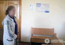 Blurred-face woman standing with hands clasped in a waiting area of a police station, beside a wooden bench, with posters on the wall and the Ukrainian National Police emblem in the corner.