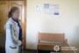 Blurred-face woman standing with hands clasped in a waiting area of a police station, beside a wooden bench, with posters on the wall and the Ukrainian National Police emblem in the corner.
