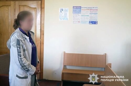 Blurred-face woman standing with hands clasped in a waiting area of a police station, beside a wooden bench, with posters on the wall and the Ukrainian National Police emblem in the corner.