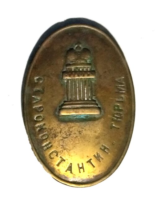 Oval brass badge featuring a central column monument relief and Cyrillic text around the edge; likely a commemorative or organizational pin.