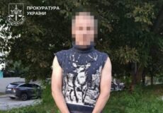 Man with hands cuffed standing on a grassy verge beside a sidewalk, wearing a sleeveless knit sweater and gray pants; face blurred; Ukrainian prosecutor logo in the corner.