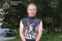 Man with hands cuffed standing on a grassy verge beside a sidewalk, wearing a sleeveless knit sweater and gray pants; face blurred; Ukrainian prosecutor logo in the corner.