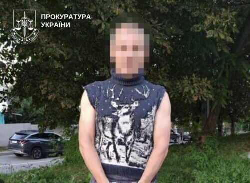 Man with hands cuffed standing on a grassy verge beside a sidewalk, wearing a sleeveless knit sweater and gray pants; face blurred; Ukrainian prosecutor logo in the corner.