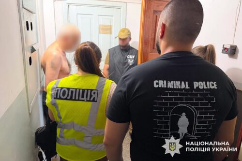 Shirtless man with a blurred face surrounded by police in a hallway; one officer wears a 'CRIMINAL POLICE' shirt.
