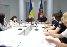 Nine people sit around a white conference table with microphones; Ukraine flag and a regional flag hang behind them.
