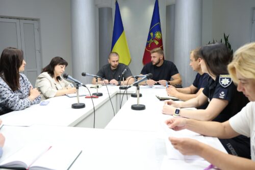 Nine people sit around a white conference table with microphones; Ukraine flag and a regional flag hang behind them.