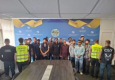 Group of people standing behind a conference table in a briefing room, several wearing neon vests labeled Migration Service in front of a blue backdrop with an emblem.