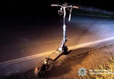 Nighttime scene of a damaged electric scooter on the roadside, police badge visible in the lower-right corner.