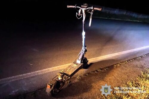 Nighttime scene of a damaged electric scooter on the roadside, police badge visible in the lower-right corner.
