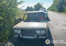 Beige sedan parked on a rural road with the driver's door open; a police vehicle is visible in the background and the Ukrainian National Police badge appears in the lower right corner.
