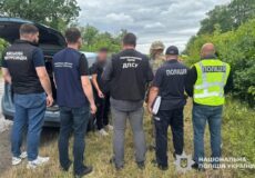 Group of Ukrainian police and security officers gathered around an open car trunk in a grassy roadside area, discussing or inspecting something. Several wear vests labeled Поліція or ДПС, with insignia visible in the background.