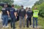 Group of Ukrainian police and security officers gathered around an open car trunk in a grassy roadside area, discussing or inspecting something. Several wear vests labeled Поліція or ДПС, with insignia visible in the background.