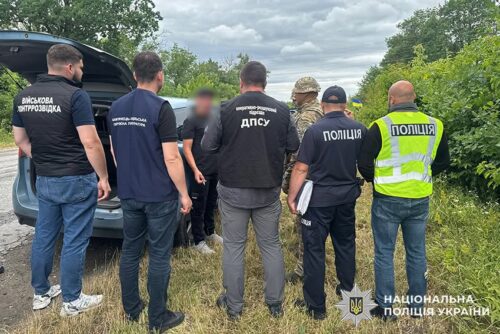 Group of Ukrainian police and security officers gathered around an open car trunk in a grassy roadside area, discussing or inspecting something. Several wear vests labeled Поліція or ДПС, with insignia visible in the background.