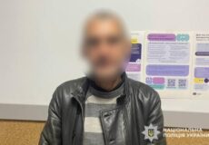 Blurry man in a black leather jacket and striped sweater standing indoors beside informational posters, with National Police of Ukraine logo.
