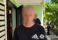 Man in a black Adidas shirt stands outside a building by a row of mailboxes; face blurred.