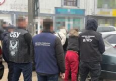 Group of men in dark jackets with SBU and прокуратура markings, standing with backs to the camera near a car by storefronts