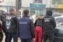 Group of men in dark jackets with SBU and прокуратура markings, standing with backs to the camera near a car by storefronts