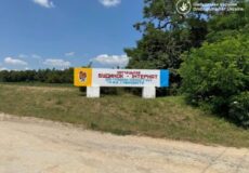 Billboard on a grassy roadside with blue and yellow panels reading 'Будинок - Інтернат' and a logo, set near trees and a dirt road.