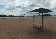 Sandy beach with blue shade structures and yellow benches, calm water in the distance under a cloudy sky