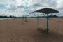 Sandy beach with blue shade structures and yellow benches, calm water in the distance under a cloudy sky