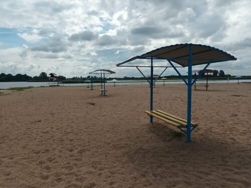 Sandy beach with blue shade structures and yellow benches, calm water in the distance under a cloudy sky