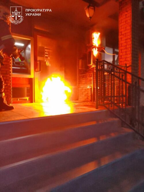 Fire erupts at the entrance of a brick building; a firefighter stands on the left as flames blaze near the doorway, with the Ukrainian Prosecutor's Office logo in the top-left corner.