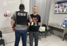 SBU officer in a bulletproof vest stands beside a man with a pixelated face inside a Ukrainian prosecutor’s office, logo visible on the wall