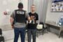 SBU officer in a bulletproof vest stands beside a man with a pixelated face inside a Ukrainian prosecutor’s office, logo visible on the wall