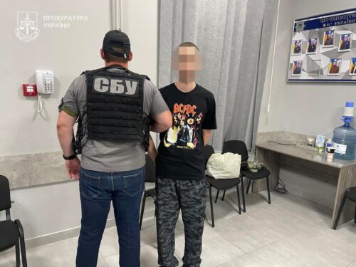 SBU officer in a bulletproof vest stands beside a man with a pixelated face inside a Ukrainian prosecutor’s office, logo visible on the wall